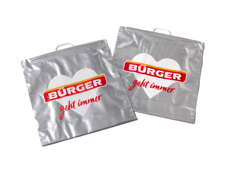 Zwei silberne Kühltaschen mit BÜRGER-Logo und Aufdruck „geht immer“ auf großem weißen Herz.