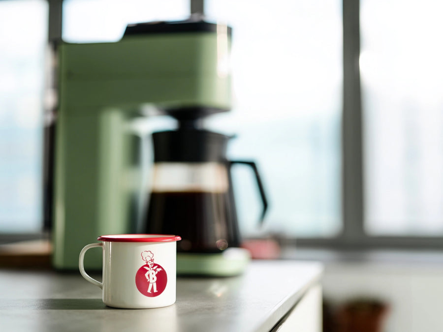Weiße BÜRGER-Tasse mit rotem Rand steht vor Kaffeemaschine in heller Küchenumgebung.