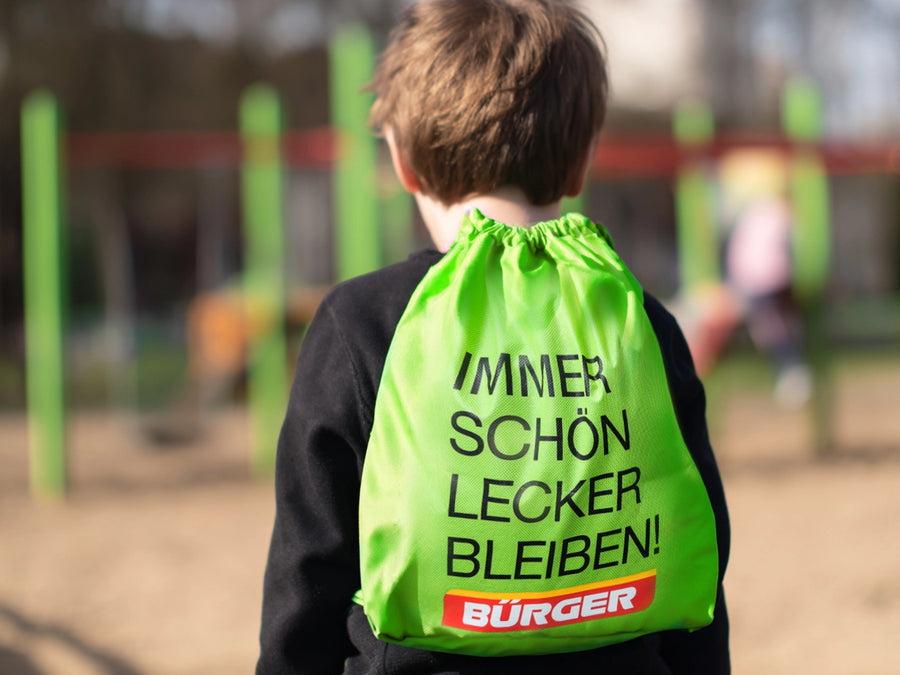 Kind mit grünem BÜRGER-Turnbeutel, Aufdruck „Immer schön lecker bleiben!“ auf dem Rücken.