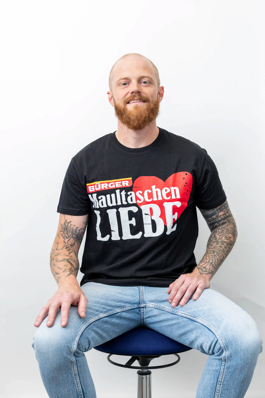 Mann sitzt auf einem Hocker mit BÜRGER-T-Shirt, rotem Herz und weißem Aufdruck „Maultaschen Liebe“.