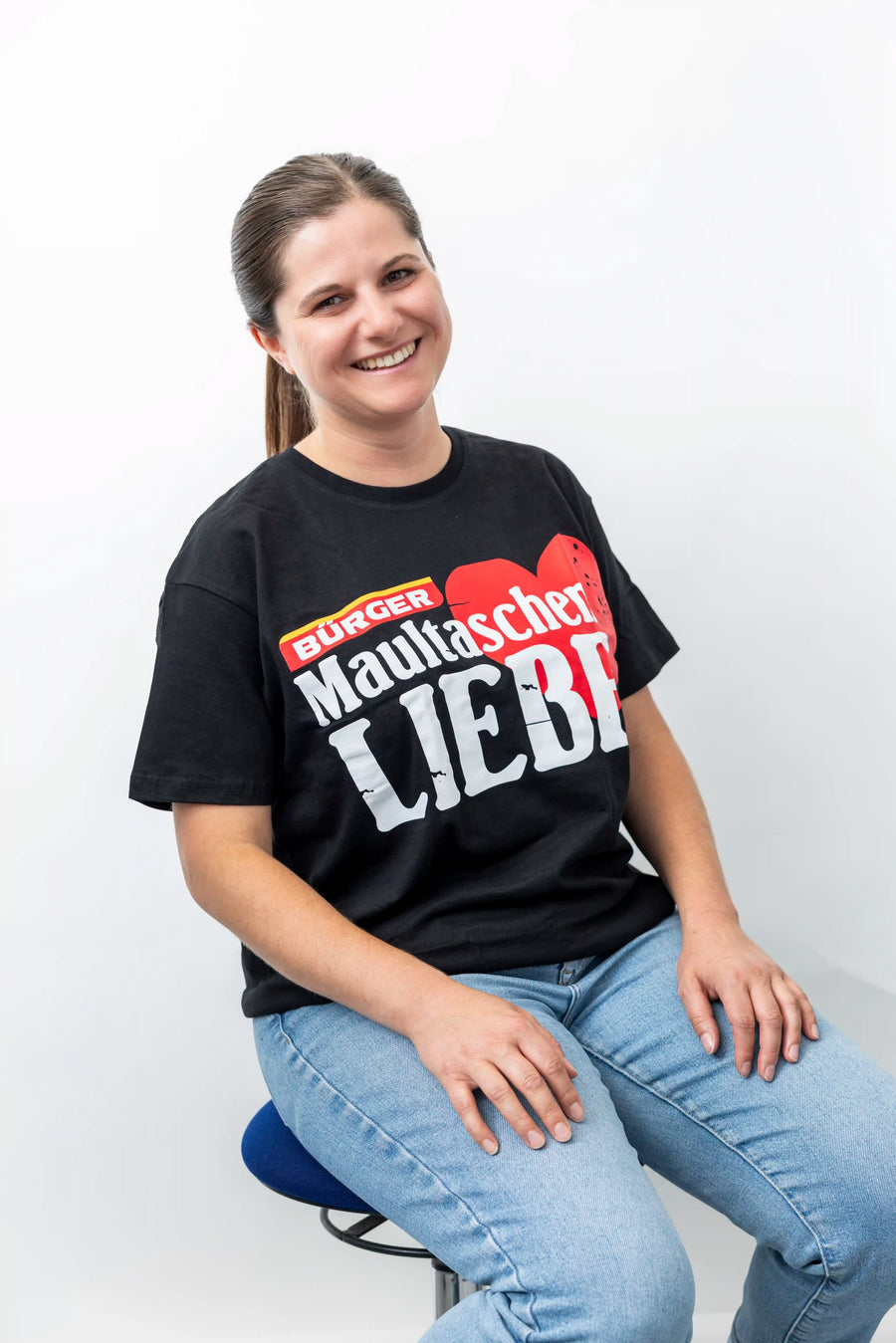 Frau sitzt und trägt ein schwarzes BÜRGER-T-Shirt mit rotem Herz und dem Aufdruck „Maultaschen Liebe“.
