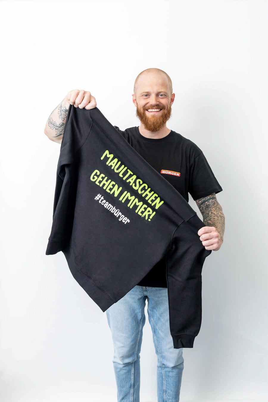 Mann mit BÜRGER-T-Shirt hält schwarzen Hoodie mit grünem Aufdruck „Maultaschen gehen immer“.