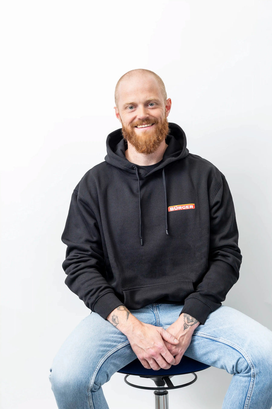 Mann trägt schwarzen BÜRGER-Hoodie mit Kapuze, sitzt auf Hocker und lächelt frontal.