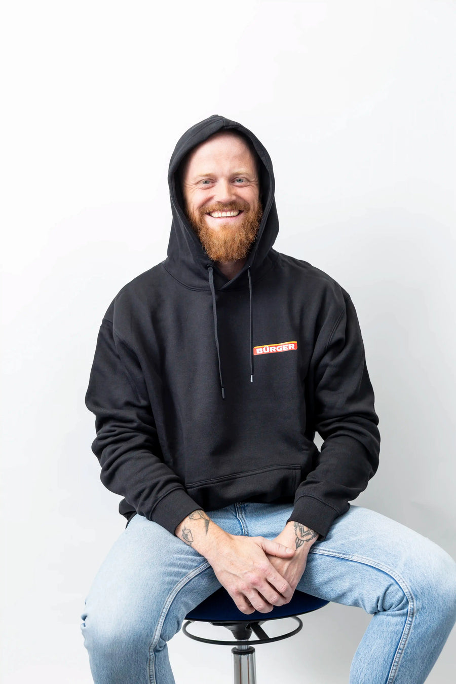 Mann mit schwarzem Hoodie von BÜRGER, Kapuze aufgesetzt, sitzend vor weißem Hintergrund.