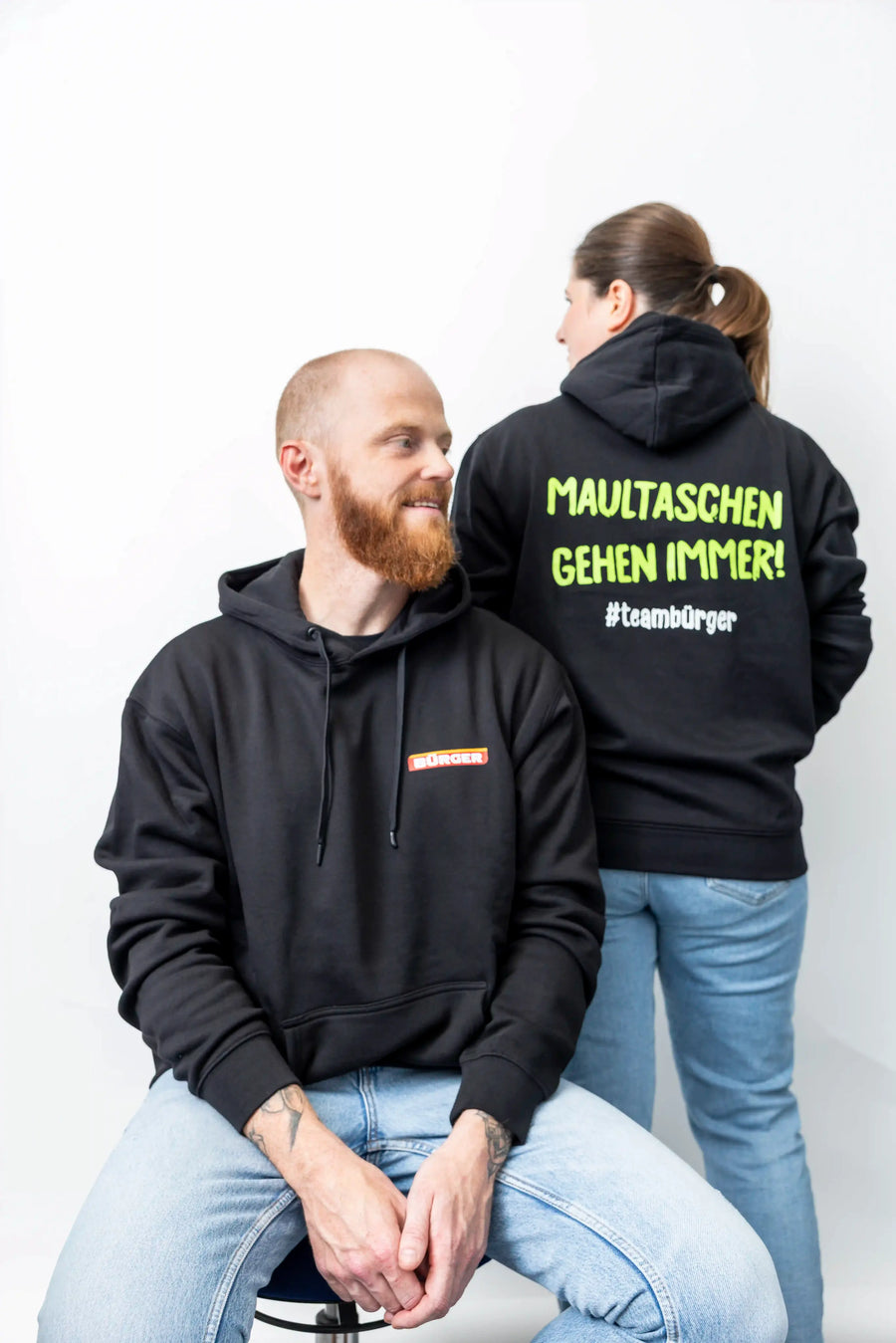 Mann sitzt im BÜRGER-Hoodie, Frau steht daneben mit Rückendruck „Maultaschen gehen immer!“.