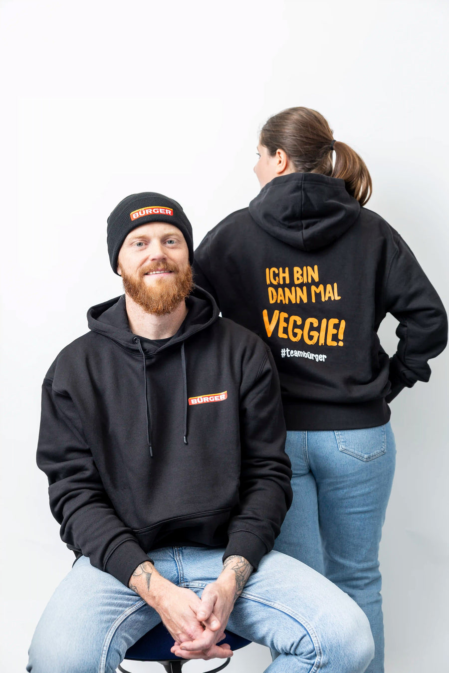 Mann mit BÜRGER-Beanie und Hoodie, Frau zeigt Rückseite mit gelbem Aufdruck „Ich bin dann mal veggie!“.
