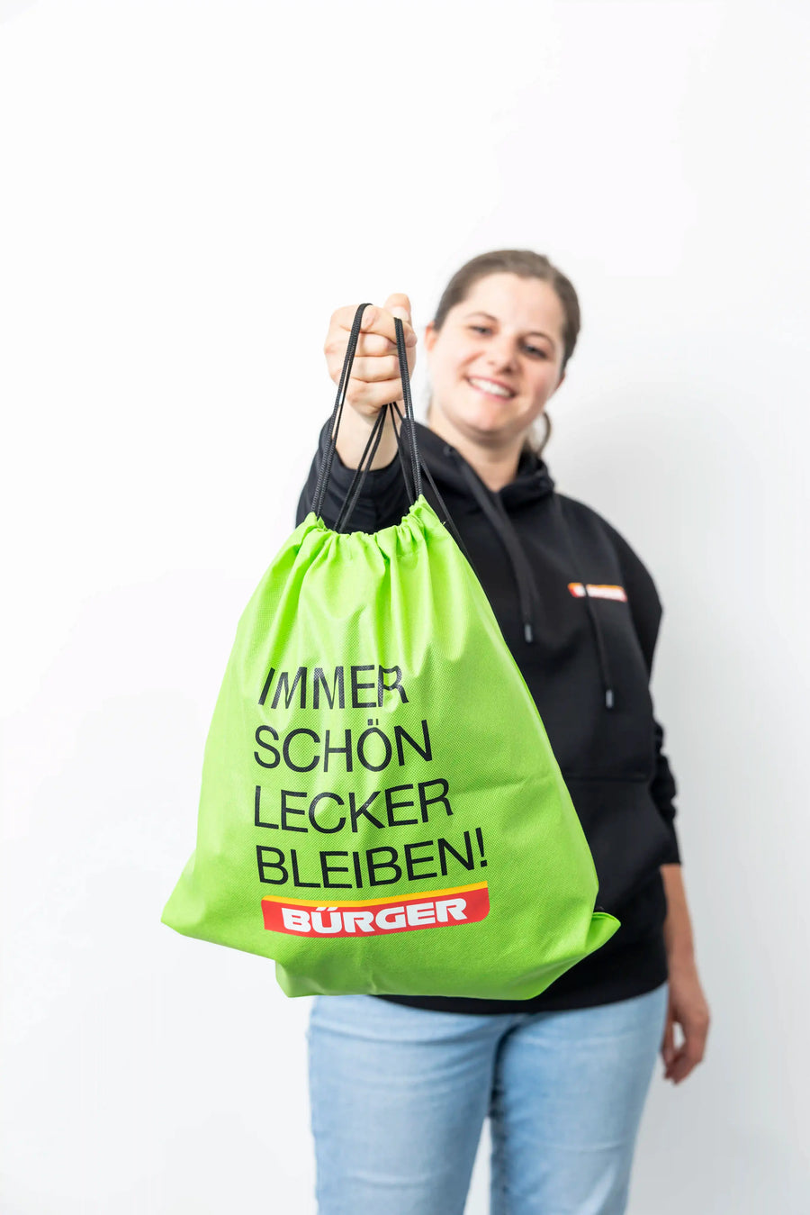 Frau hält grünen Turnbeutel mit schwarzem Aufdruck „Immer schön lecker bleiben!“ und BÜRGER-Logo.