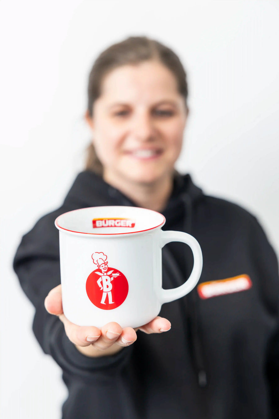 Frau hält weiße Emaille-Tasse mit BÜRGER-Logo und gezeichnetem Koch, Vordergrund scharf gestellt.