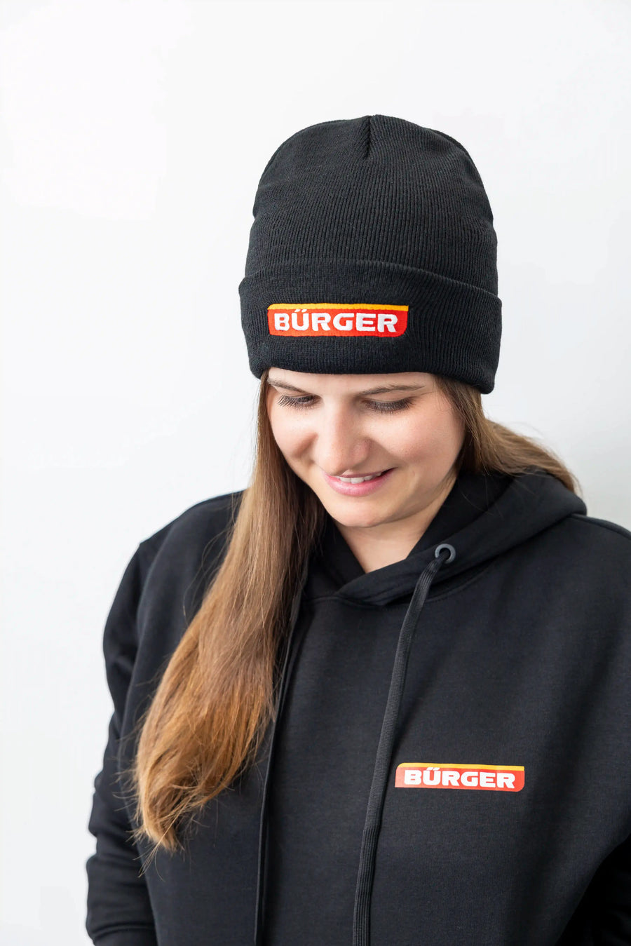 Frau mit schwarzer BÜRGER-Mütze und Hoodie, blickt nach unten und lächelt leicht.