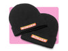 Zwei schwarze BÜRGER-Beanies mit gesticktem Logo auf rosa Hintergrund nebeneinander.